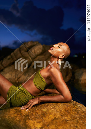 Trans sexual black fashion model in open dress, brass jewelry poses lying on big stone inside natural pool surrouded by rocks on tropical island at night. Androgynous ethnic person in backwater. 113974042