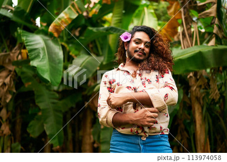 South Asian gay man with floral shirt, blue jeans stands in tropical foliage, purple flower in hair. Confident smile, celebrates inclusivity, pride, LGBT identity amidst natures embrace. South Asian gay man with floral shirt, blue jeans stands in tropical foliage, purple flower in hair. Confident smile, celebrates inclusivity, pride, LGBT identity amidst natures embrace. 113974058