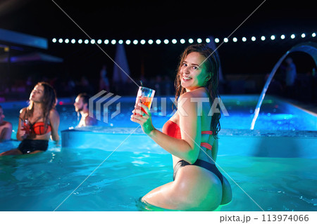 Pretty woman in bikini drinking cocktail and hanging out with friends at night pool party. People having fun dancing with drinks and splashing water in luxury private villa swimming pool. 113974066