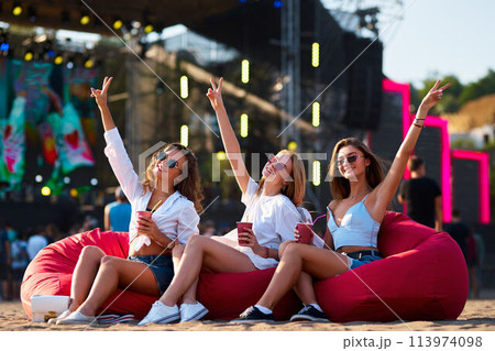 Group of joyful girls on red bean bags at sunset, cheering with drinks, enjoying live music at sandy beach fest. Casual summer outfit, sunglasses, vibrant festival vibes, ocean backdrop. Group of joyful girls on red bean bags at sunset, cheering with drinks, enjoying live music at sandy beach fest. Casual summer outfit, sunglasses, vibrant festival vibes, ocean backdrop. 113974098