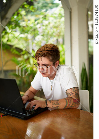 Transgender entrepreneur in casual wear works on laptop at eco-friendly tropical coworking space. Focused on digital tasks, promotes startup culture, values diversity. Green surroundings. 113974130