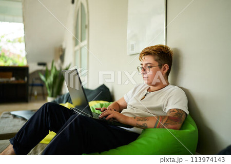 Transgender pro works on laptop from cozy bean bag in modern coworking space. Focused on screen, person enjoys flexible working environment. Inclusive workplace, remote job and casual office setup. Transgender pro works on laptop from cozy bean bag in modern coworking space. Focused on screen, person enjoys flexible working environment. Inclusive workplace, remote job and casual office setup. 113974143