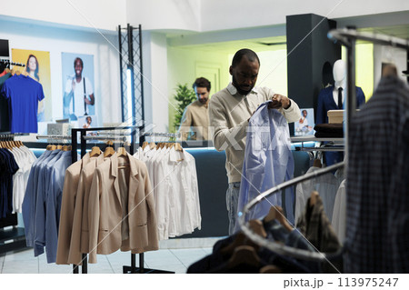 African american buyer checking size on shirt label while choosing trendy formal wear in shopping mall boutique. Young man browsing fashionable apparel on hanger in showroom 113975247