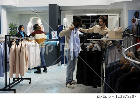 Clothing store customer trying on shirt and getting advice from boutique employee. African american man holding apparel to body while choosing size with fashion outlet assistant Clothing store customer trying on shirt and getting advice from boutique employee. African american man holding apparel to body while choosing size with fashion outlet assistant 113975287