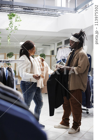 Boyfriend offering girlfriend to try on casual jacket in clothing store. Shopping mall woman customer holding trendy blazer on hanger and asking man friend for advice before buying 113975502
