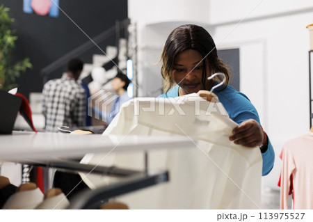 African american customer holding hanger with white shirt, checking items fabric in modern boutique. Shopaholic woman looking at new fashion collection, shopping for casual wear in clothing store 113975527