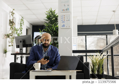 Business arab professional texting on smartphone while working on laptop in start up office. Entrepreneur searching financial information on mobile phone in green open space Business arab professional texting on smartphone while working on laptop in start up office. Entrepreneur searching financial information on mobile phone in green open space 113975566