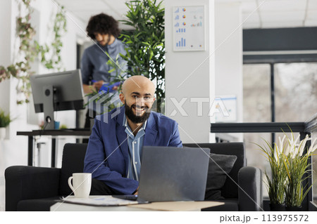 Smiling business entrepreneur working on laptop and looking at camera in start up company office. Successful project manager using portable computer while sitting on couch portrait 113975572