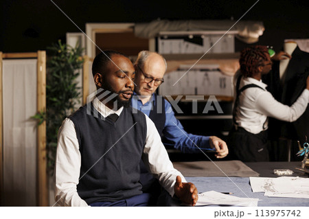 Master tailor taking client measurements while meticulous seamstress apprentice does needlework on suit in background. Gentleman getting custom made attire done by professional couturiers team 113975742