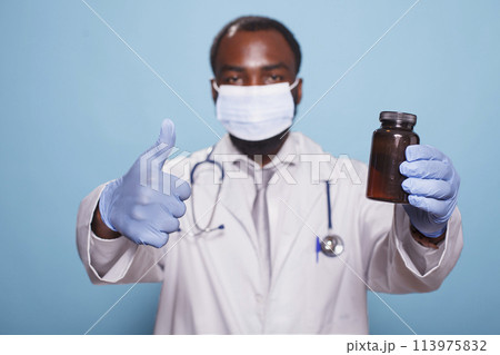 Portrait of medical specialist in a face mask, showing thumbs up while gripping pills bottle. Black doctor with stethoscope and gloves approving prescription painkillers with hand gestures. Portrait of medical specialist in a face mask, showing thumbs up while gripping pills bottle. Black doctor with stethoscope and gloves approving prescription painkillers with hand gestures. 113975832