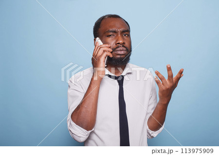 Frowning male freelancer explaining problem on smartphone doing hand gesture against blue background. Upset african american businessman in bad mood trying to understand business conversation. 113975909