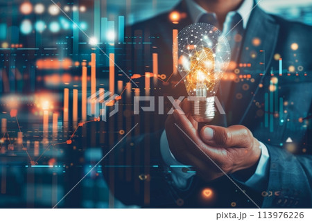 Businessman's hand holding a glowing light bulb in front of financial data charts in a close-up shot 113976226