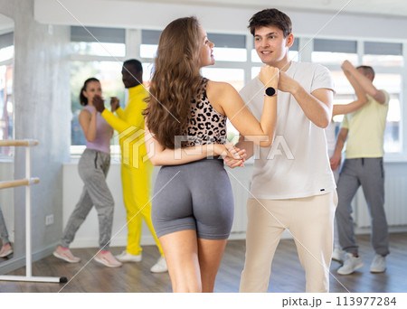 Smiling woman and man in pair enjoying dance and training movements Latin dance bachata with multinational group active people in dancing class Smiling woman and man in pair enjoying dance and training movements Latin dance bachata with multinational group active people in dancing class 113977284