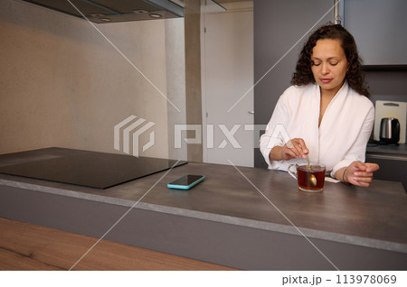 Beautiful curly brunette in white bathrobe, standing at kitchen counter and adding sugar to hot drink. Charming young woman stirring sugar in a cup of tea while having a breakfast in the home kitchen. Beautiful curly brunette in white bathrobe, standing at kitchen counter and adding sugar to hot drink. Charming young woman stirring sugar in a cup of tea while having a breakfast in the home kitchen. 113978069