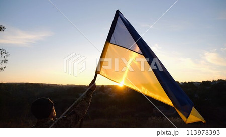 Male soldier stands with raised flag of Ukraine against background of beautiful sunset. Male ukrainian army soldier with a lifted blue-yellow banner in honor of the victory against russian aggression Male soldier stands with raised flag of Ukraine against background of beautiful sunset. Male ukrainian army soldier with a lifted blue-yellow banner in honor of the victory against russian aggression 113978393