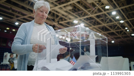 Elderly female voter puts bulletin into box, looks at camera Elderly female voter puts bulletin into box, looks at camera 113991303