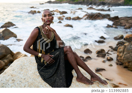 Non-binary ethnic fashion model in dress, brass jewelry sits on rocks by ocean. Trans sexual black person with rings, nose-ring, bracelets, earrings in posh clothes poses in tropical seaside location. 113992885