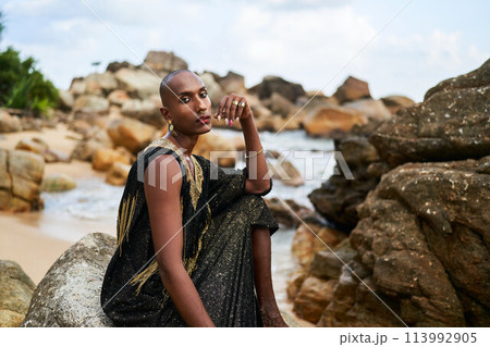 Non-binary ethnic fashion model in luxury dress sits on rocks by ocean. Trans sexual black person in jewellery and posh clothes poses gracefully in tropical seaside location. Pride month. 113992905