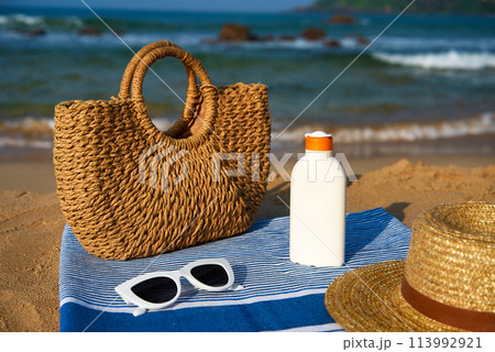 Beach essentials on sandy shore woven bag, white sunscreen bottle, stylish sunglasses atop blue towel, straw hat alongside. Waves crash near summer seaside accessories for sunny day on coast. Beach essentials on sandy shore woven bag, white sunscreen bottle, stylish sunglasses atop blue towel, straw hat alongside. Waves crash near summer seaside accessories for sunny day on coast. 113992921