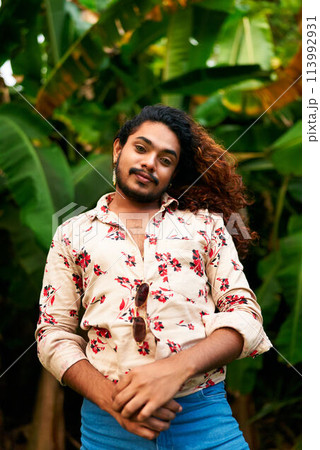 Confident gay man with curly hair stands in tropical setting. Flamboyant pose, bright floral shirt open, denim pants. Arms crossed, smiling, gender expression, lush green foliage backdrop. 113992931
