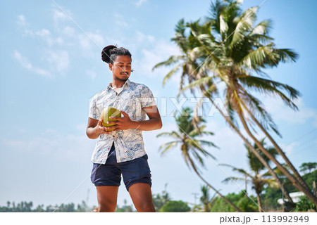 Bipoc man enjoys fresh coconut on sunny tropical beach. Male quenches thirst with natural drink, relaxes by palm trees, embodies laid-back vacation vibe, explores wellness, leisure travel trend. Bipoc man enjoys fresh coconut on sunny tropical beach. Male quenches thirst with natural drink, relaxes by palm trees, embodies laid-back vacation vibe, explores wellness, leisure travel trend. 113992949