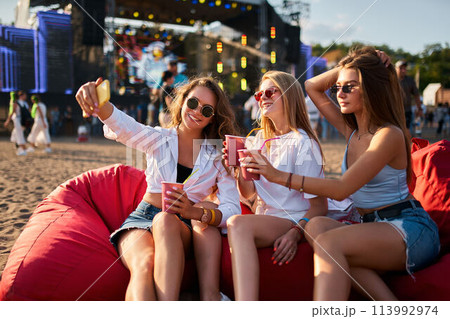 Group of women enjoy music fest on beach, lounging on bean bags. Casual attire, sunglasses, sipping drinks, one takes selfie. Warm daylight highlights relaxed, fun atmosphere of outdoor summer event. Group of women enjoy music fest on beach, lounging on bean bags. Casual attire, sunglasses, sipping drinks, one takes selfie. Warm daylight highlights relaxed, fun atmosphere of outdoor summer event. 113992974