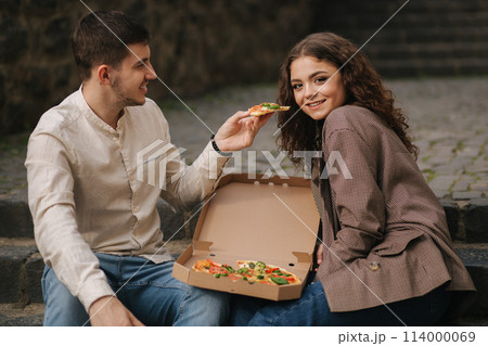 Smiling love couple eating vegan pizza at street on stairs. Handsome man give slice of pizza to woman 114000069