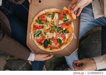 Top view image of couple grab slices of pizza from box at the outdoor. Man and woman hands taking pizza. Vegan pizza with fresh tomatoes basil and broccoli. Lactose and gluten free 114000072