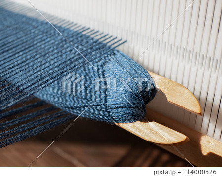 Wooden weaving shuttle with blue yarn is inserted in open shed. Separation between upper and lower warp yarns through which the weft is woven 114000326