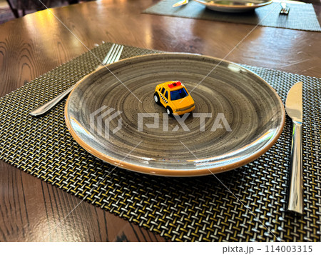 Miniature taxi toy on ceramic plate with cutlery. Concept of calling taxi after end of banquet or celebration. Design of taxi advertising, culinary presentations and unusual concepts. 114003315