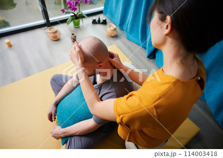 Masseuse making thai yoga massage. Woman therapist doing traditional massage treatment for male patient indoors. Bald client sitting on mat, meditating, enjoying hand massage in calm atmosphere. 114003418