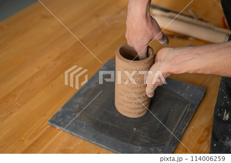 Close-up of a man's hands making a patterned cylinder out of clay.  114006259