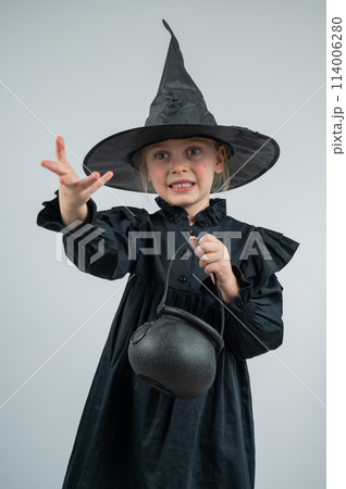 Portrait of a little Caucasian girl in a witch costume holding a cauldron on a white background. Vertical photo.  114006280