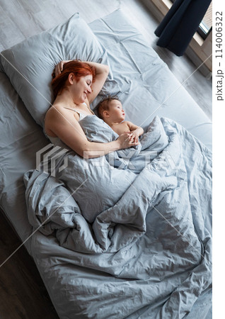 Top view of a red-haired Caucasian woman lying in bed with her baby son. Vertical photo. Top view of a red-haired Caucasian woman lying in bed with her baby son. Vertical photo. 114006322