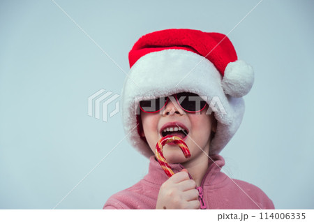 Portrait of a cute Caucasian girl in a Santa Claus hat and sunglasses eating a lollipop on a white background.  114006335