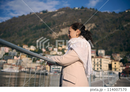 Side portrait pensive serene woman standing on the lakefront, admiring beautiful Como lake in Lombardy. People and travel concept. Mesmerizing view of Italian Alps mountains on backdrop on sunny day Side portrait pensive serene woman standing on the lakefront, admiring beautiful Como lake in Lombardy. People and travel concept. Mesmerizing view of Italian Alps mountains on backdrop on sunny day 114007570