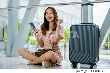 A female traveler sitting on the ground with a suitcase and smartphone, contemplating her journey. She enjoys the peaceful solitude of travel. Adventure is her escape. 114009739