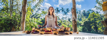 Woman playing on Tibetan singing bowl while sitting on yoga mat against a waterfall. Vintage tonned. Beautiful girl with mala beads meditating BANNER, LONG FORMAT 114011305