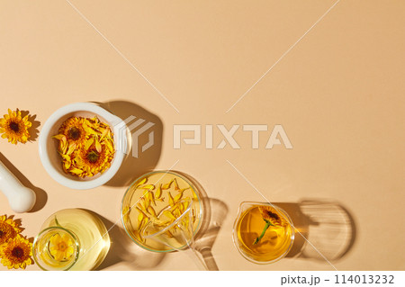 Laboratory glassware filled with liquid and oil extracted from Calendula flowers. A white mortar containing yellow flowers and petals featured. Vacant space for product advertising. Copy space 114013232