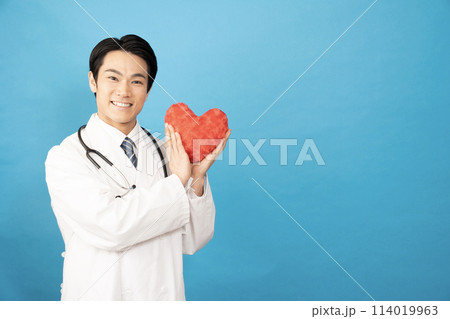 ブルーバックで赤いハートを持って微笑む男性医師の写真素材  