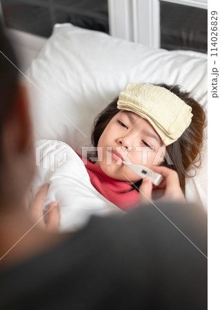 Asian mother measuring temperature girl with digital thermometer in her mouth on bed at morning time, Sick child have cool towel for reduce high fever, Selective focus, Healthy and infection concept Asian mother measuring temperature girl with digital thermometer in her mouth on bed at morning time, Sick child have cool towel for reduce high fever, Selective focus, Healthy and infection concept 114026829