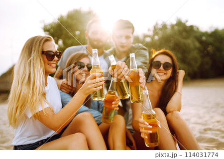 Group of friends cheers and drink beers on the beach. Young friends relaxing and having picnic. Suummertime. 114031774
