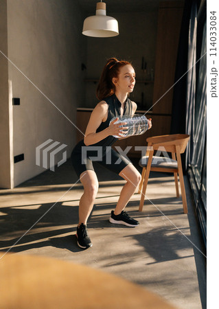 Vertical shot of sporty woman doing squat exercise holding in hands bottle with water. Athletic fit female wearing sportswear crouching training muscles workout at home for healthy body by window. Vertical shot of sporty woman doing squat exercise holding in hands bottle with water. Athletic fit female wearing sportswear crouching training muscles workout at home for healthy body by window. 114033084