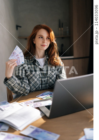 Vertical shot of happy businesswoman holding banknotes of dollars in and fanning money in front of face. Successful female enjoying prosperity sitting at laptop with remote computer at home office. 114033096