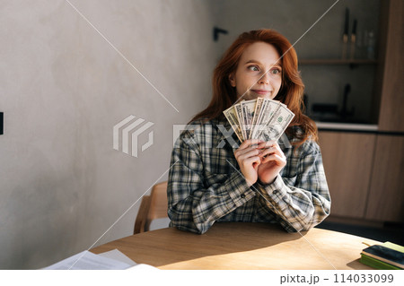 Funny fooling young woman counting money in dollars banknotes sitting in kitchen table at home. Rich happy female holding in hands stack of cash money of success, gain, victory. Concept of investment. 114033099