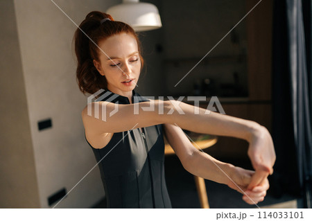 Portrait of athletic young woman in outfit preforming warm-up exercise, stretching arm muscles before workout indoors, standing by window with on sunny day. Athlete prepare muscle for exercise workout 114033101