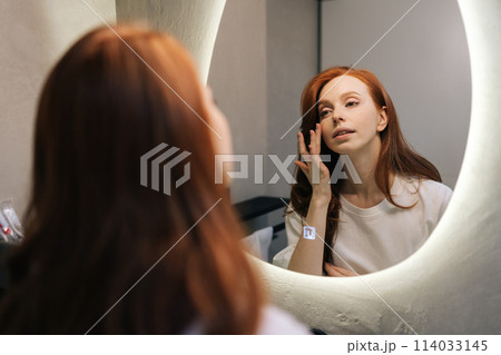 Reflection on mirror of attractive young redhead woman enjoying her appearance while getting ready in bathroom. Happy beautiful female touching face look in bathroom mirror, enjoy healthy skin. 114033145