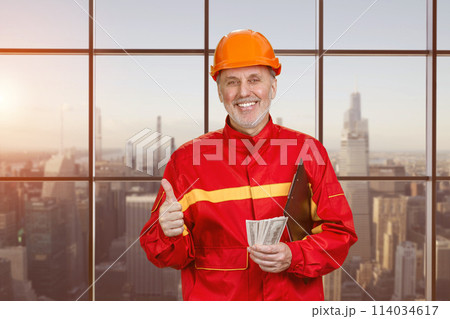 Portrait of happy elder man holding money and showing like gesture. WIndows with view of urban cityscape background. 114034617