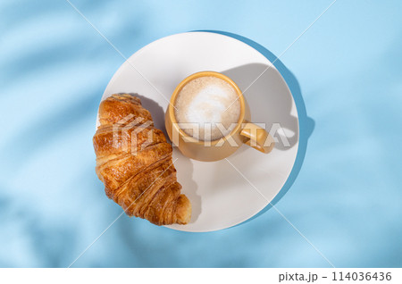 Flat lay of coffee cup and fresh croissant on a white plate on a blue background with shadow. Flat lay of coffee cup and fresh croissant on a white plate on a blue background with shadow. 114036436