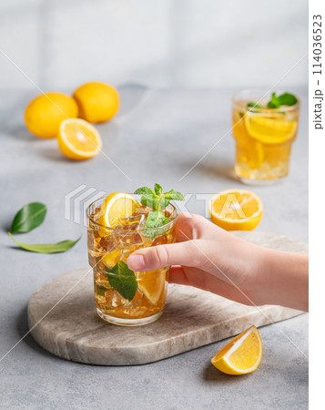 A hand takes a glass of iced tea with lemon and mint against a gray background with citrus fruits 114036523
